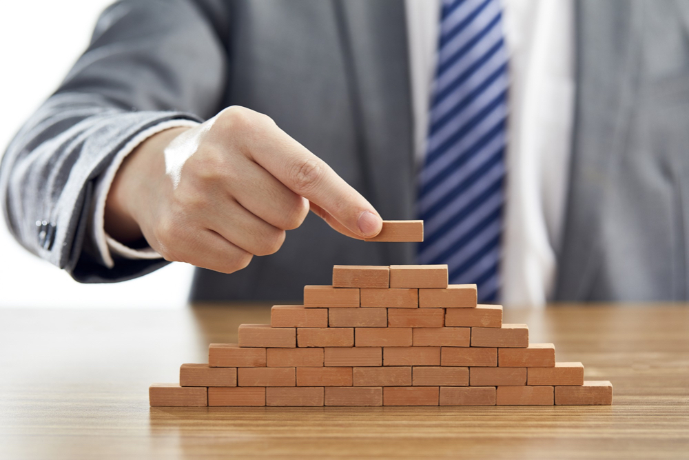A business man in formal suit, putting small pieces of wooden blocks forming a pyramid, in representation of an enterprise SEO business growth campaign.