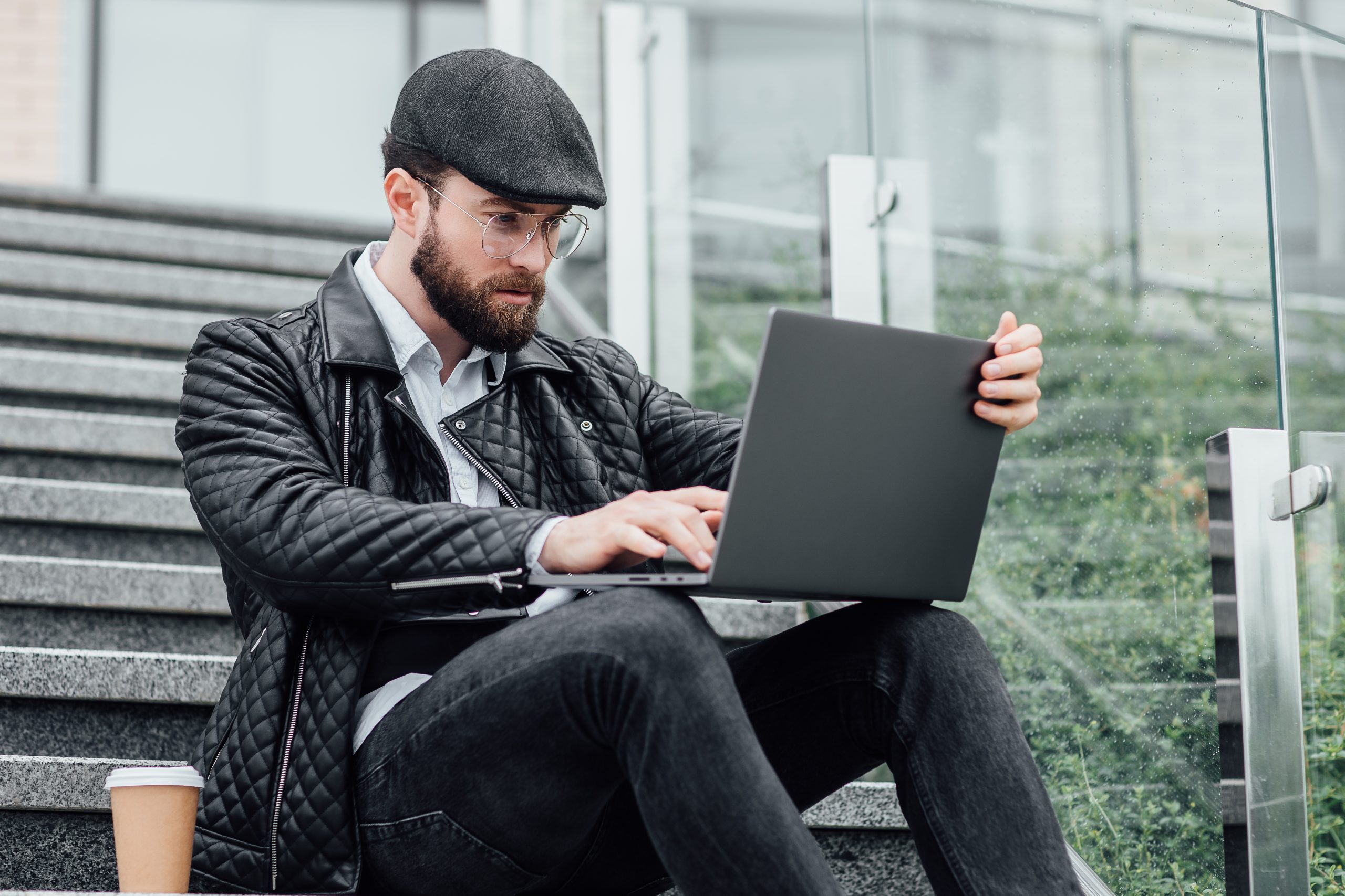 A young man on a black hat, sitting on a stair while checking his laptop
