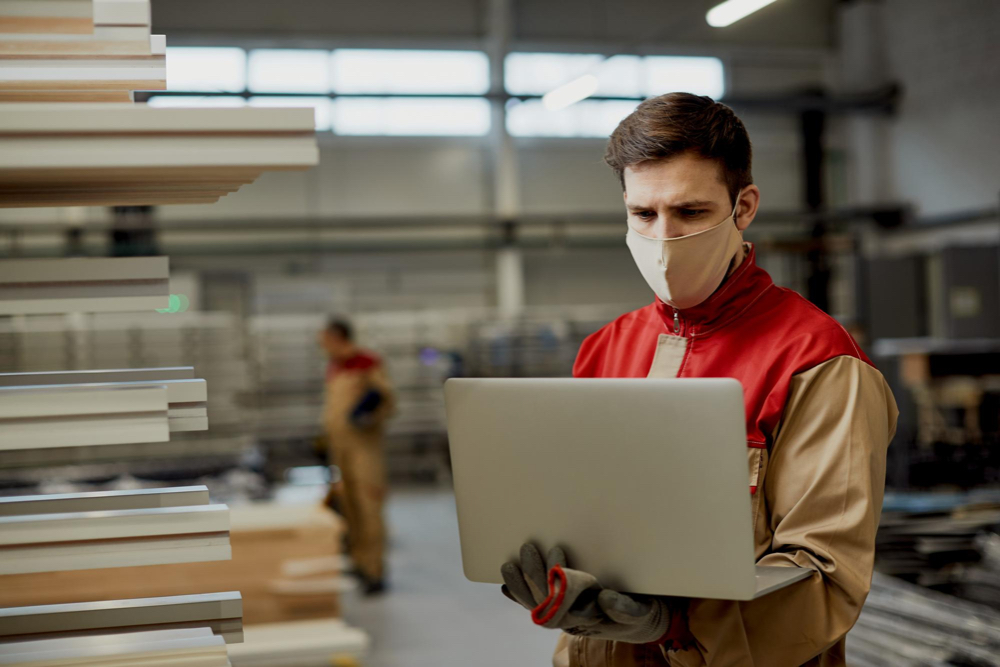 A carpentry business owner in full suit and masked on, working on his local SEO for carpentry business strategy, using a laptop, inside his warehouse.