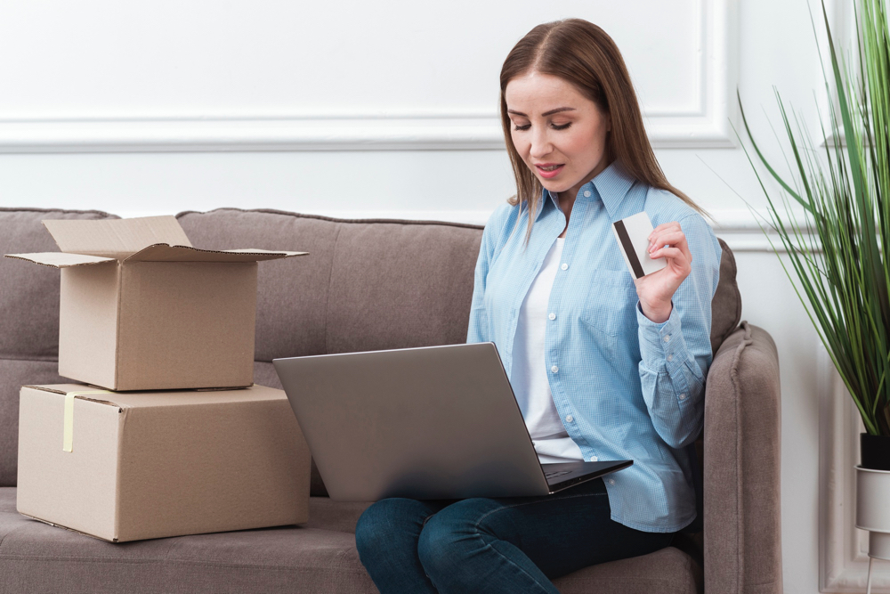 A woman doing some online shopping in a D2C business website using a laptop, with a card at hand and shopping boxes at the side of the sofa.