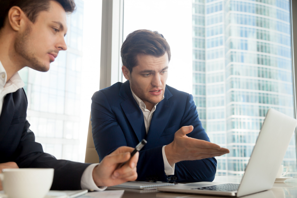 Two digital marketers in formal business suits inside an office board room, one explaining a technical SEO audit report, the other one's taking dow notes.