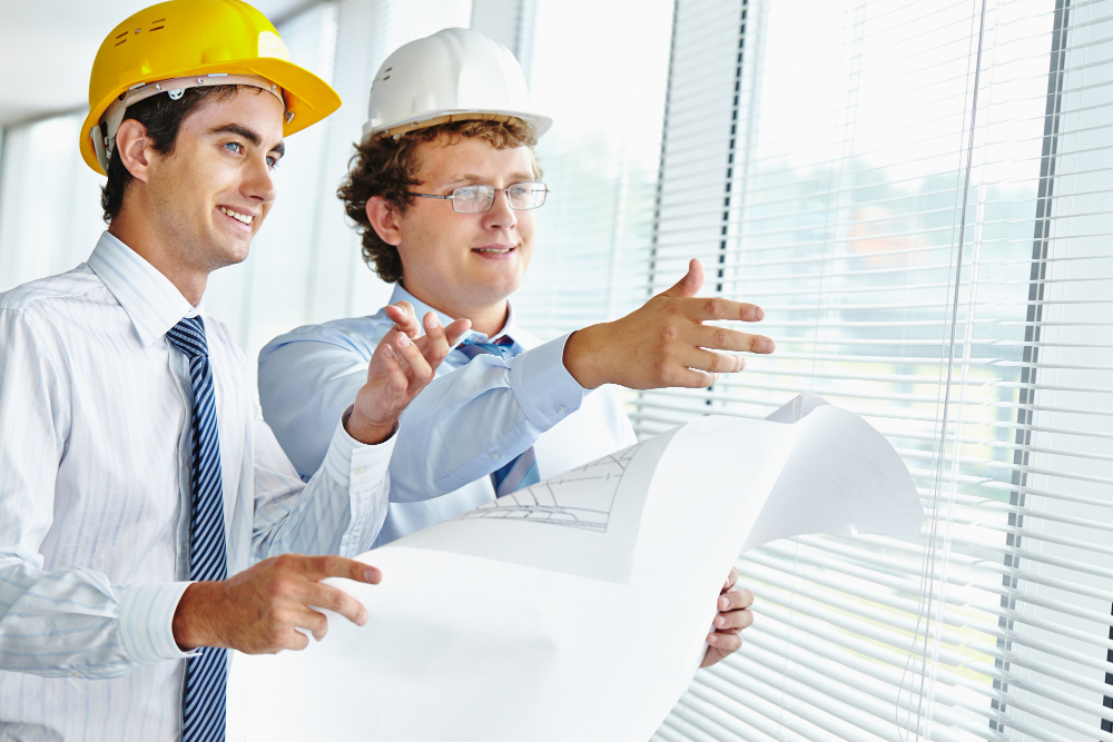 Two engineers in casual business suits, looking at a construction site while holding a drawing plan.