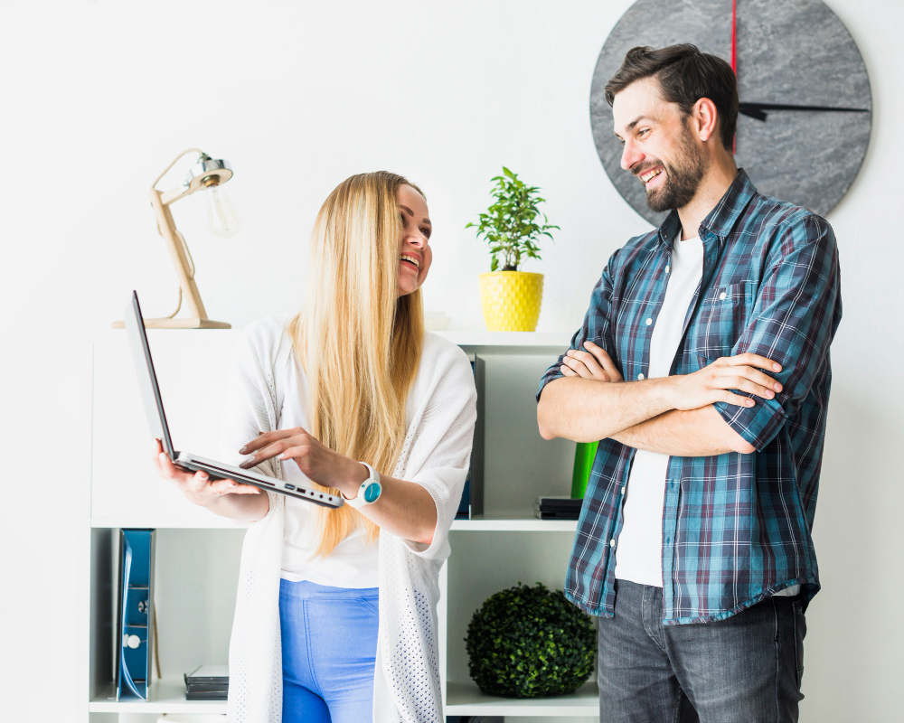 Two cheerful interior designers discussing about home & decor renovations, one holding the laptop, the other one arms crossed, smiling and listening.