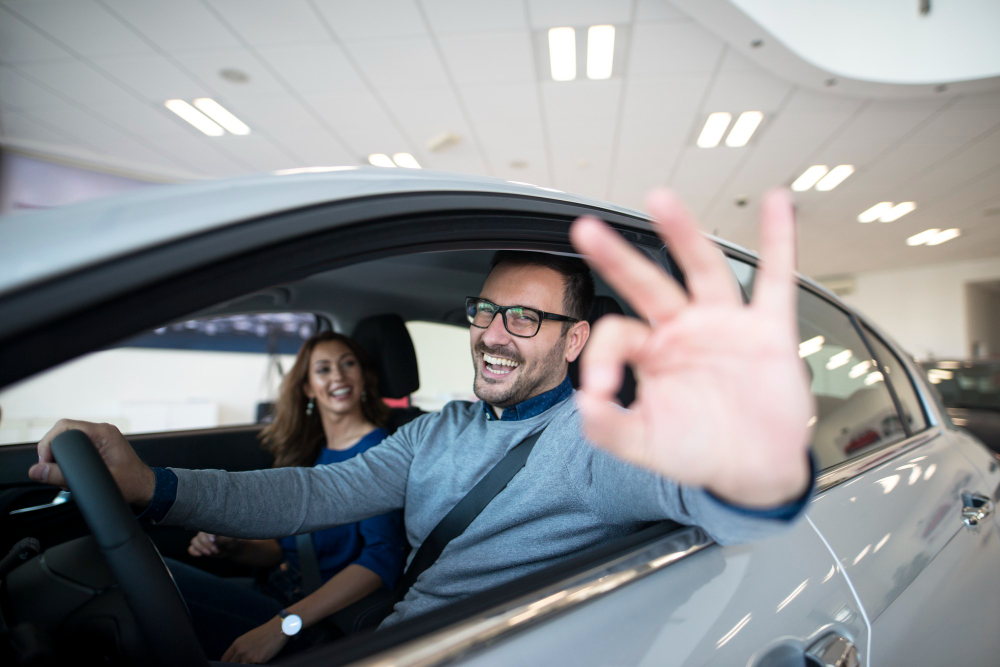 A happy customer riding a newly bought car, doing an ok sign, with his wife inside.