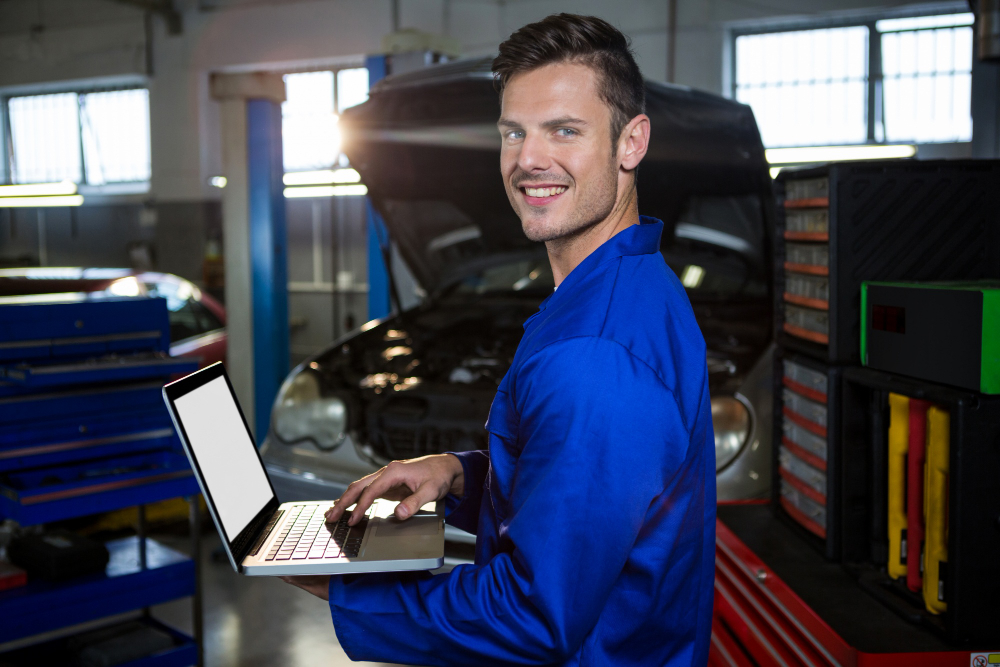 A happy mechanic working on his laptop, inside his automotive shop.
