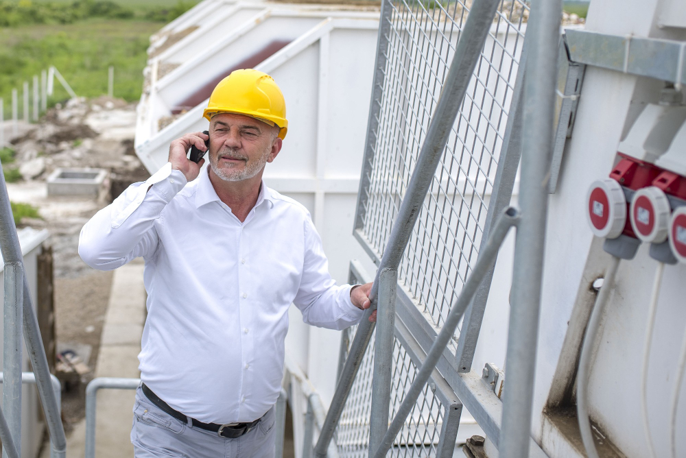 An engineer in casual white long sleeve polo and yellow hard hat, making a phone call on the side of a construction site.
