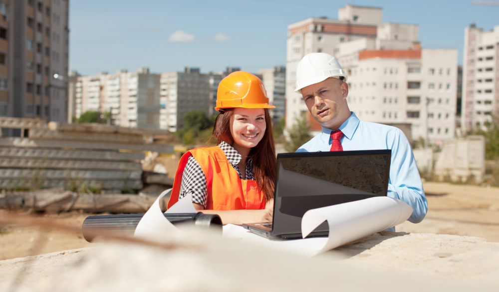 Two engineers in casual business suit and hard hats, posing for a shot on a construction site, with a laptop and drawing plan on top of a table.