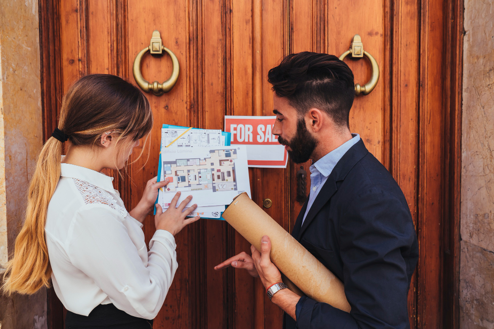 Two real estate agents in casual business suits, checking out a house plan, in front of a huge wooden door.