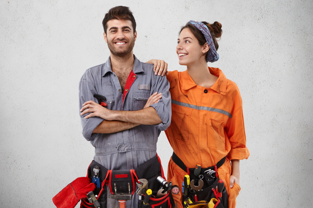 Two plumbers in jumpsuits, posing for a shot with some tools on their belly,