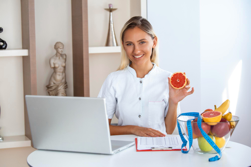 A nutritionist holding a citrus orange, post for a shot inside an office space, with a laptop and fruit basket at the front of the table.